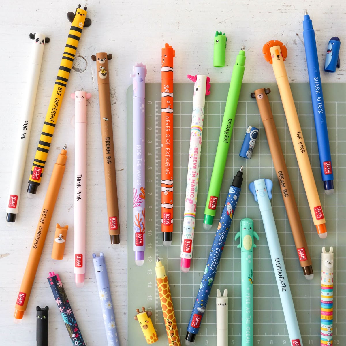 A collection of cute, animal-themed mechanical pencils arranged on a green cutting mat over a white wooden surface. The pencils feature various animal designs like a tiger, bee, panda, bear, frog, giraffe, and rabbit, with some showing visible 'ZIG' branding.